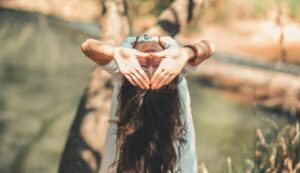 Une femme aux longs cheveux effectuant une posture de yoga près d’une rivière paisible, dans un cadre extérieur ensoleillé.