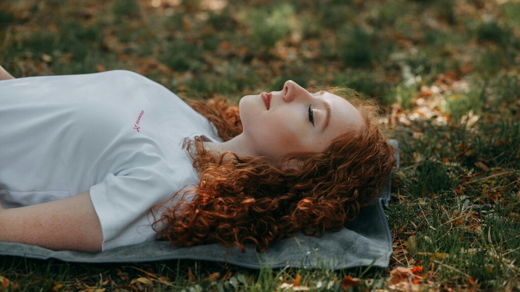 Adult woman with red hair resting peacefully outdoors on a sunny day, surrounded by nature.