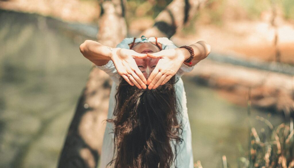 A woman with long hair performing a yoga pose by a tranquil river in a sunny outdoor setting.